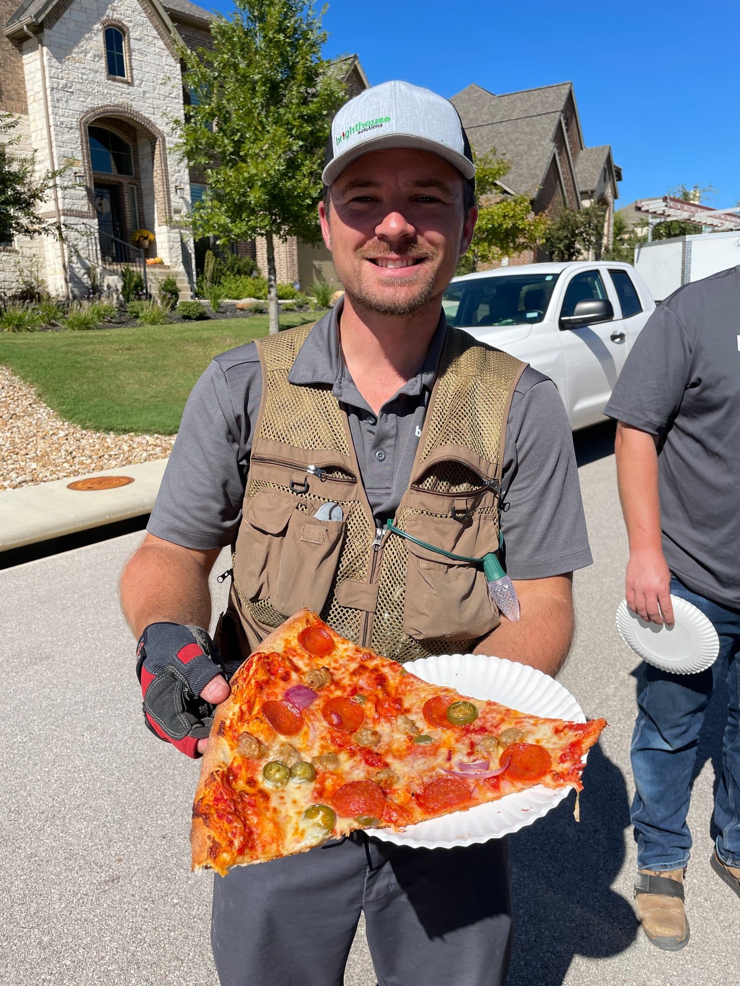 A smiling installer in a work vest holding up an enormous slice of pepperoni pizza on a paper plate.