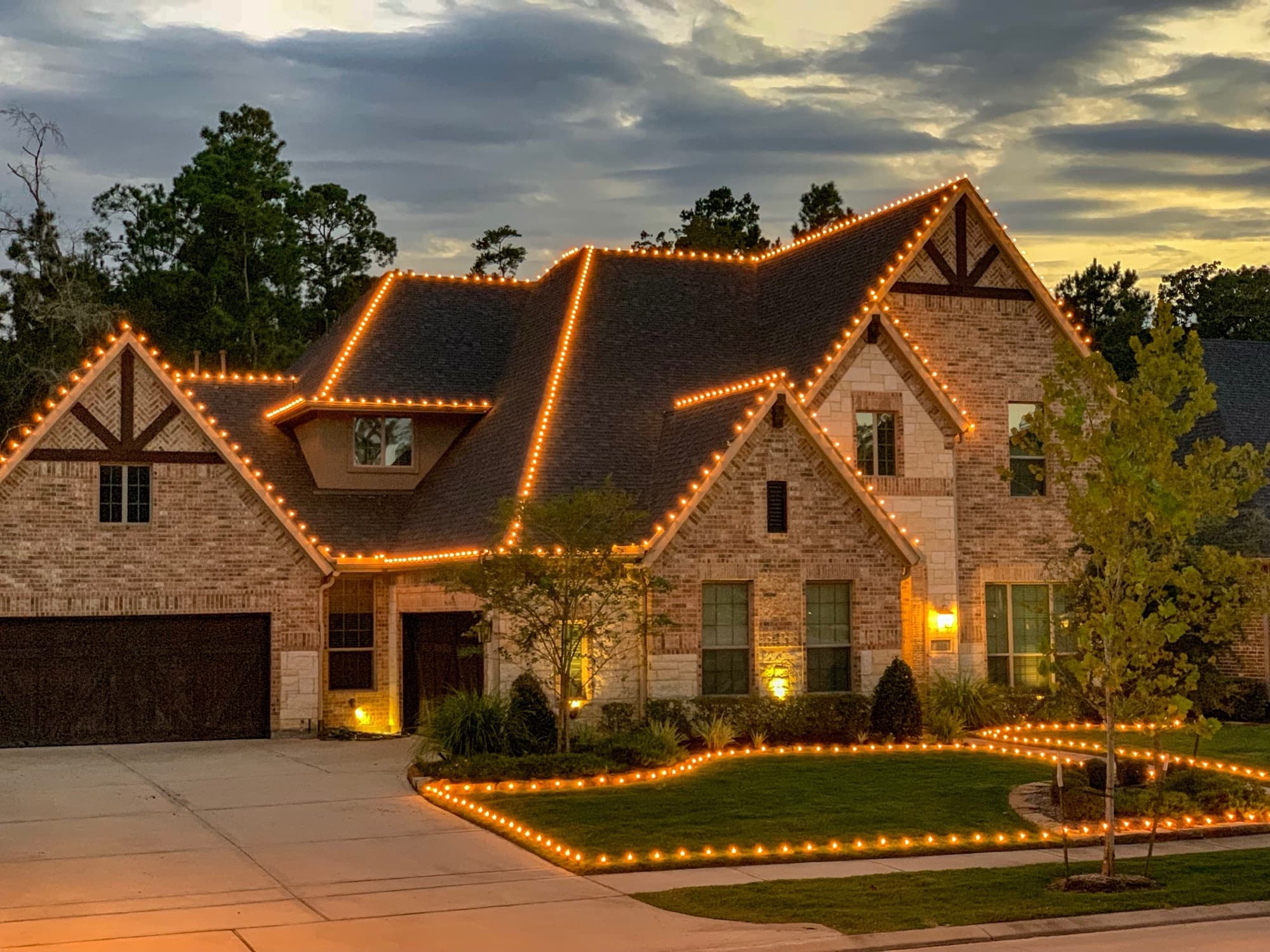 A brick two-story home at dusk, every roofline and flowerbed traced in warm white Christmas lights.