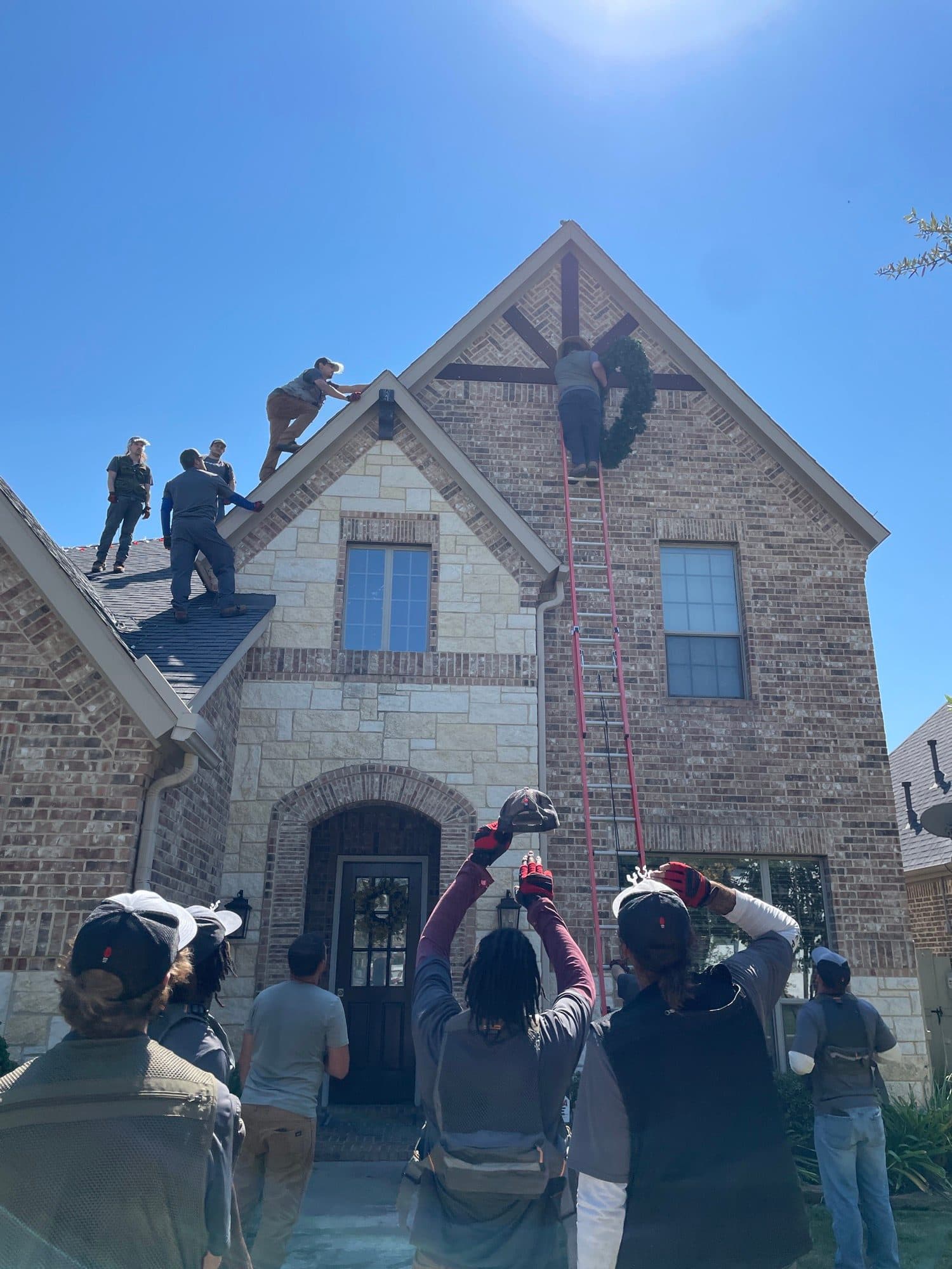 A full crew gathered in front of a brick home during preseason bootcamp, watching a wreath-hanging demonstration on a ladder two stories up.