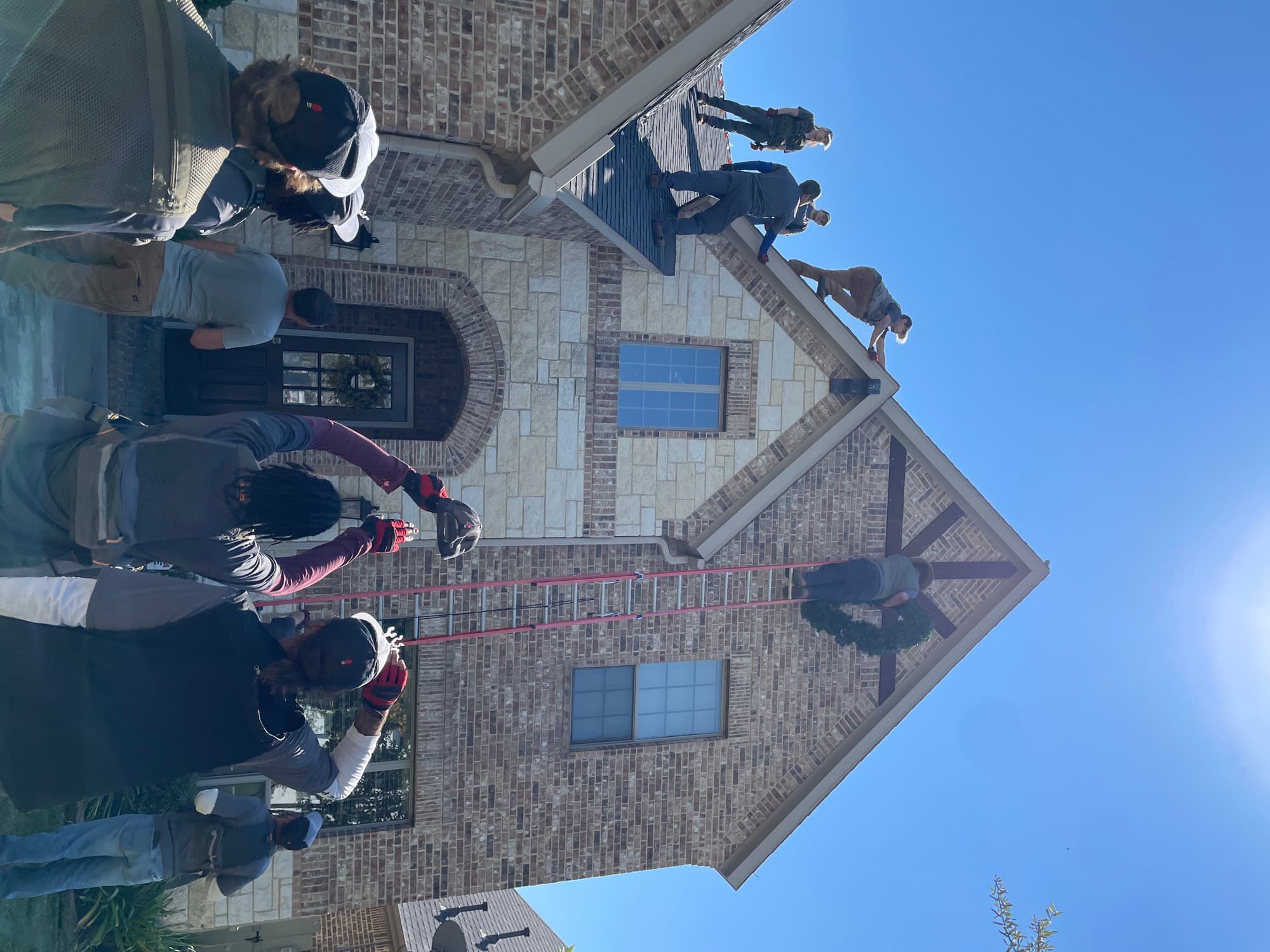 A full crew gathered in front of a brick home during preseason bootcamp, watching a wreath-hanging demonstration on a ladder two stories up.