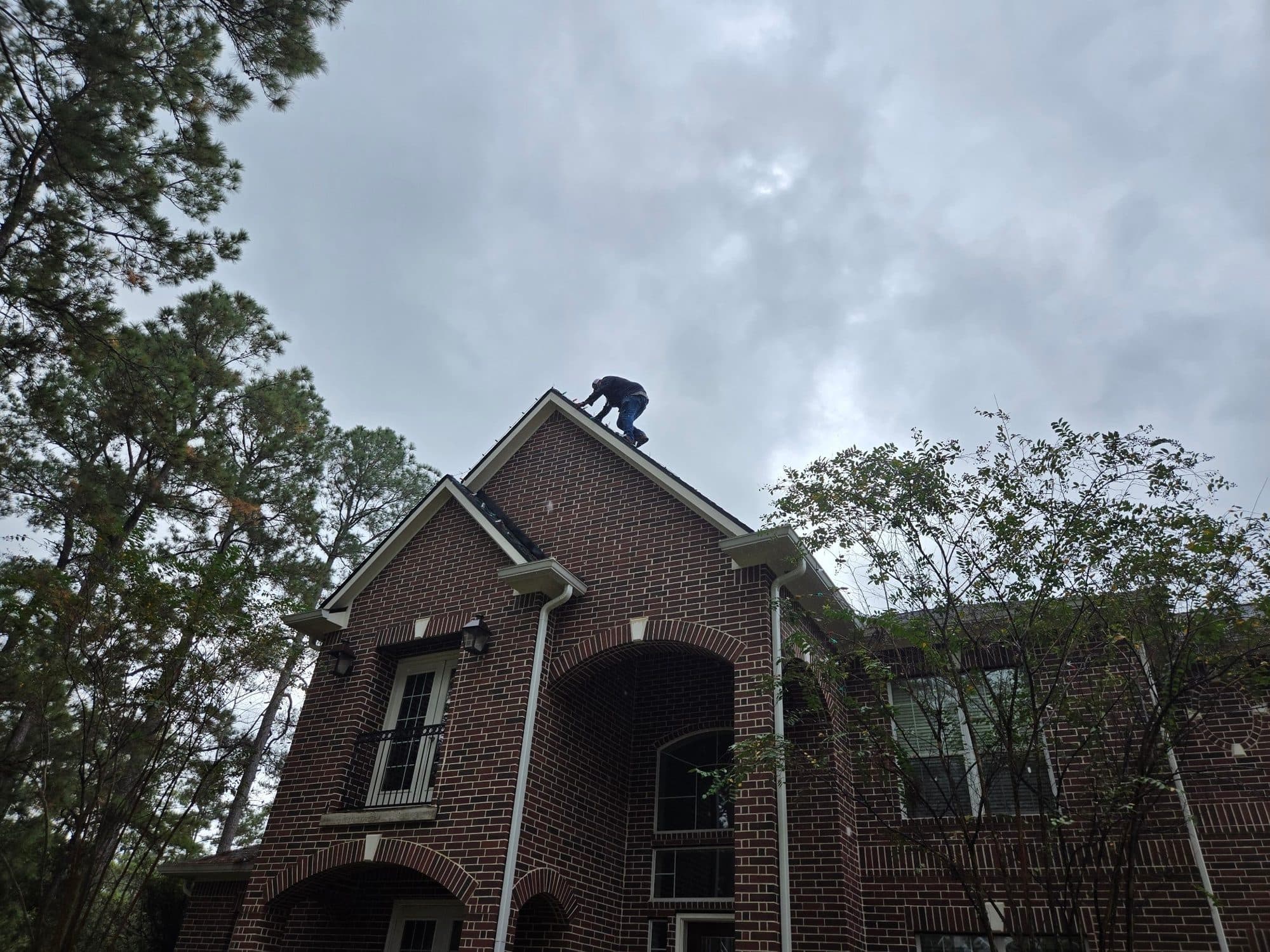 An installer working along a steep roof ridge under a gray sky, hanging Christmas lights.