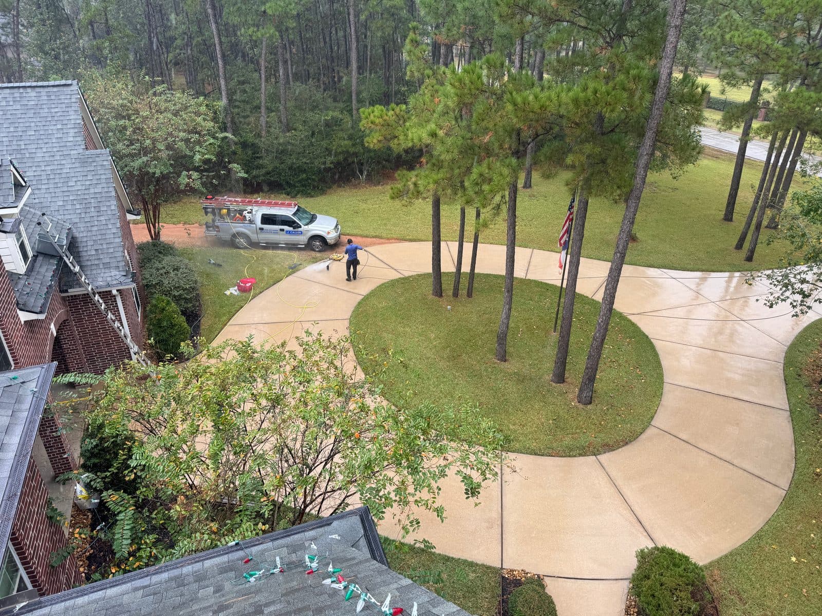 Aerial view of a technician running a surface cleaner across a large circular driveway with a pine island, work truck parked on the lawn behind him.